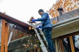 Man in ladder removing autumn leaves from gutter during day of autumn